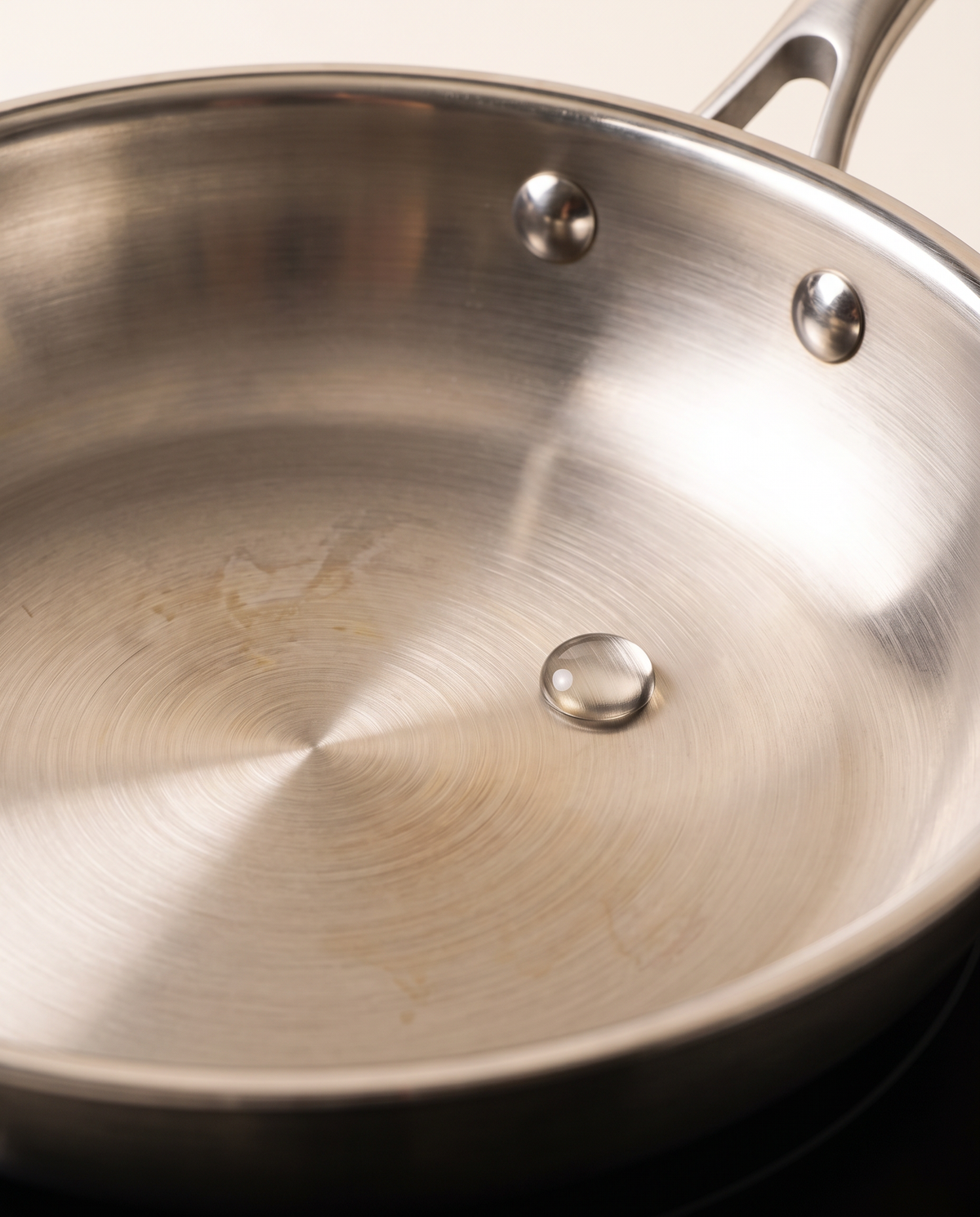 Une goutte d'eau perlée en bille ronde qui roule sur le fond brossé d'une poêle inox Marthe préchauffée, les deux rivets du manche visibles sur la paroi